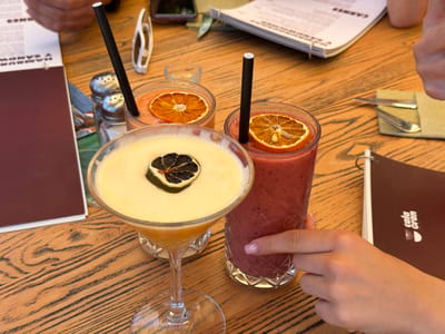The image captures a moment at a table in what appears to be a cafe or restaurant in Cala D'or, Spain, as suggested by the 'cala gran' logo on a menu. The table is made of light-colored wood with dark grain. Three drinks are placed on the table: a cocktail in a stemmed glass with a dried lime garnish, a bright orange drink in a short glass with an orange slice garnish, and a thick red smoothie in a tall glass with an orange slice garnish. All the drinks have black straws. A hand is touching the glass of the smoothie. Beside the drinks, there are menus, salt and pepper shakers, sunglasses, and a set of cutlery wrapped in a napkin. Additional hands and parts of arms suggest that there are other people present.