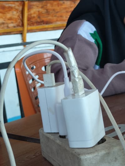 The image depicts two white USB wall chargers plugged into a worn power strip on a wooden table. The chargers are connected to white USB cables. One of the cables appears to be reinforced with clear tape near the connector. A portion of a person is visible in the background, wearing a dark head covering and a taupe-colored sleeve with a white and green decorative element. An orange plastic chair is partially visible behind the chargers. The image was reportedly taken in Matang, Indonesia.