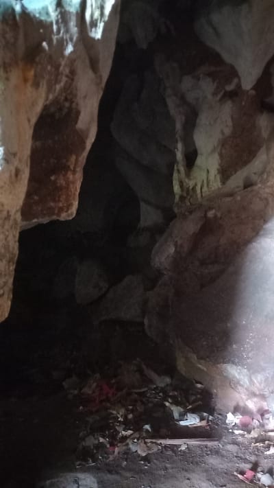 The image appears to be taken inside a cave in Thap Put, Thailand. The foreground features rough, light-colored rock formations with patches of brown. A dark, seemingly bottomless cavity is visible behind and between the rocks. The upper right area of the image shows a section of the cave floor covered in what seems to be debris, including leaves, pieces of cloth (some of which are red), and other miscellaneous litter. A wooden plank is also visible amidst the debris. The lighting within the cave is dim and uneven. There are no people visible.