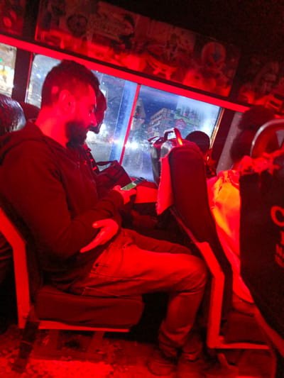 The image displays the interior of a public transport vehicle, likely a Kenyan matatu, during nighttime or evening hours. The setting is characterized by a dominant red artificial lighting that bathes the entire cabin, creating a distinct atmosphere.

In the left foreground, a male passenger with a beard, wearing a dark hoodie and jeans, is seated and intently looking down at a mobile phone he holds in his hands. His left arm is casually folded across his body. Behind him, another passenger is partially visible, obscured by shadow. To the right, in the seat ahead, a person wearing a yellow garment is seen from the back. The seats are dark, and what appear to be bags or luggage are visible near the passengers' feet and next to the main subject.

Through the vehicle's window, an urban or suburban street scene can be observed. Buildings and distant artificial lights illuminate the outside environment. On one of the buildings visible through the window, fragmented text "KFS [unclear number]M" can be discerned. The upper interior of the vehicle features various posters or decorations, also illuminated by the red light.

The activity depicted is that of passengers commuting, with at least one person engaging with a mobile device. The strong red lighting inside contrasts with the cooler tones of the exterior lights. On the far right, near the back of a seat or a passenger's clothing, a white letter "C" is faintly visible. The scene suggests typical public transport in the Kahawa Sukari ward of Kenya.