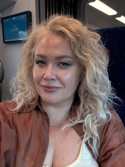 A woman with blonde, wavy hair is captured in a close-up, angled shot. She is wearing a brown button-up shirt over a light-colored top. Her gaze is directed slightly away from the camera, and she has a gentle smile.
The setting appears to be inside a train or public transport, suggested by the blue upholstered seating and what looks like a window or light fixture in the background. A digital screen is visible in the lower left of the frame, displaying text. The text is in Hebrew and reads "תל אביב" (Tel Aviv) and "רכבת ישראל" (Israel Railways), indicating the location is likely within Israel. The overall lighting suggests it is daytime. The emotional tone of the image is relaxed and candid.