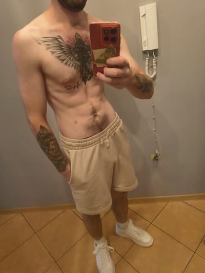 A shirtless person with tattoos on their chest and left arm is standing in a room, taking a selfie with a phone. The person is wearing light-colored shorts and white sneakers. The room has a grey wall with a white intercom device and a tiled floor. The image does not provide any information about the location being Grójec, Poland.
