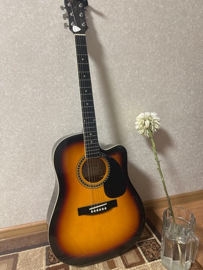 An acoustic guitar with a sunburst finish leans against a textured, beige wall. To the right of the guitar, a single, dried white flower in a clear glass vase with water stands on a wooden floor. The guitar's headstock is visible at the top of the frame, featuring tuning pegs and a white pick. The body of the guitar displays a black pickguard and six strings. A patterned rug is visible on the floor in the foreground. The lighting suggests an indoor setting, likely a room in a residential building. There are no people or animals present in the image. This scene is a still life composition.