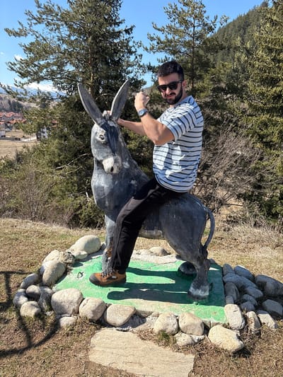 An adult male subject is depicted seated astride a weathered, dark grey donkey statue, located on a grassy, sloped area in Madzhare, Bulgaria. The subject, with dark hair and a beard, wears sunglasses, a light blue and white horizontally striped short-sleeved t-shirt, dark trousers, and brown hiking boots. He has a wristwatch on his left wrist. His right arm is bent at the elbow with a clenched fist raised, while his left hand rests on the statue's neck. The donkey statue is mounted on a green-painted rectangular base, which is bordered by irregularly shaped natural rocks. The immediate ground is dry, light brown soil with sparse green vegetation. In the mid-ground, a village with multiple houses featuring brown roofs is visible. The background consists of numerous green pine trees and bare deciduous trees, with distant snow-capped mountains under a clear blue sky. The scene is illuminated by bright daylight, casting defined shadows.