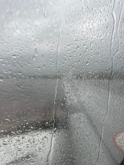 The image shows a close-up view of a window covered in raindrops. The background is out of focus and appears to be an outdoor environment with muted colors, suggesting it might be a cloudy or rainy day. The focus is entirely on the water droplets on the glass, which are of various sizes and shapes, creating a textured and somewhat abstract surface. Some droplets are elongated and appear to be running down the windowpane, while others are more rounded. The light reflects off the water, adding to the visual texture. No people, specific objects, or discernible city features are visible in detail due to the obstruction and focus. The location in Jhiurheri, India, cannot be identified from the visual information provided.