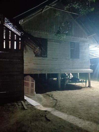 The image depicts an outdoor scene at night, likely in Asanbideun, Indonesia, featuring traditional stilt houses and surrounding ground. A stilt house, constructed from light-colored wooden planks, dominates the right side of the frame. It has a sloped roof covered with corrugated metal sheeting and two dark-colored windows with louvred shutters. The house is raised on green-painted stilts, with storage or objects visible beneath it, including what appears to be a green metal frame, possibly a bed or bench, with some items on it.

To the left of the stilt house is a darker structure, also wooden, with vertical slats that allow light to pass through from the interior, suggesting it might be another dwelling or a covered area. A small, light-colored wooden gate with vertical planks stands ajar between the two structures, leading to a pathway.

The ground is primarily dirt and sand, with patches of dry grass and a few scattered debris. Shadows are cast by the structures and a tree with foliage visible in the upper right, indicating artificial lighting from an unseen source. The overall atmosphere is dark and somewhat rustic. There are no people or animals visible in the image.