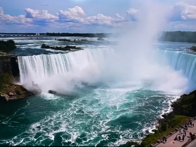 The image depicts a wide view of Niagara Falls, with the Horseshoe Falls dominating the center and right of the frame. The water is a striking turquoise color and turbulent, with white foam swirling on the surface. Massive amounts of mist rise from the falls, obscuring part of the view on the right. On the left side of the falls, the water cascades over a rocky ledge. In the foreground, on the right bank of the falls, a paved pathway runs along the edge, with a group of people observing the scene. The pathway is lined with lush green trees and rocky terrain. In the distance, beyond the falls and mist, a line of trees forms the horizon, with a bridge and some structures visible further back. The sky is a mix of blue with scattered white clouds. The scene is not located in the city of Ləcət, Azerbaijan.