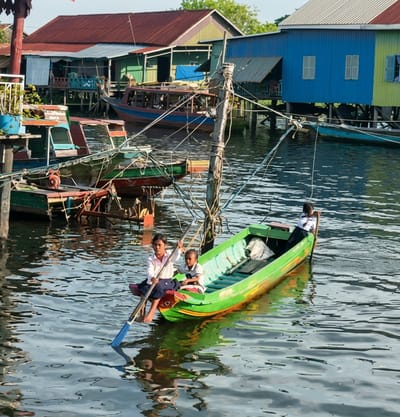 In the city of Sotr Nikom, Cambodia, a vibrant scene unfolds on a body of water with stilted houses and boats in the background. In the foreground, a bright green and yellow boat navigates the water. Three individuals are present in the boat. A young girl, wearing a white top and dark skirt, sits in the front, holding a long oar with a blue blade. To her right, a young boy in a light-colored shirt and darker shorts is seated. Behind them, another person, appearing to be a young girl, is seated in the rear of the boat, also holding an oar. The boat is surrounded by other vessels and structures, with the stilted buildings of Sotr Nikom providing a colorful backdrop of red, blue, and yellow. The water reflects the surrounding colors and the bright sunlight.