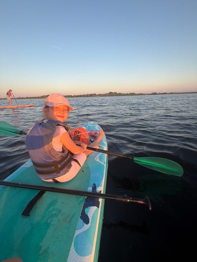 A young girl wearing a pink cap and a blue life vest sits on a paddleboard on a body of water. She is looking at the camera and smiling. The paddleboard is teal and has a blue design on it. A green paddle lies across the board. In the background, another person is paddleboarding on a bright orange board. The sky is a clear blue, and the water is calm. Trees and buildings are visible on the distant shore. The scene appears to be from Salaspils, Latvia, given the presence of paddleboarders on the Daugava River.