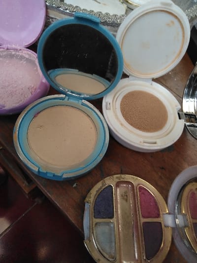 The image depicts an indoor scene with a collection of open cosmetic containers resting on a wooden surface. While stated to be located in Ciudad de San José, Costa Rica, no visual cues within the image corroborate this geographic location.
In the upper left, a light purple, circular compact holds a quantity of finely milled pinkish powder. Centrally, a round, open compact with a light blue rim features a circular mirror that reflects a portion of a beige pressed powder compact situated directly below it. To the right of the mirrored compact, an open white compact reveals a round, tan-colored cushion foundation sponge. In the lower part of the frame, a gold-toned, ornate compact is open, displaying four distinct eyeshadow pans: dark blue/purple, light blue/silver, magenta/pink, and dark purple/plum. This gold compact exhibits signs of wear, including chipped edges and surface discoloration. The items are arranged on a wooden surface, likely a table, characterized by visible grain and minor imperfections. Portions of other reflective objects or containers are visible at the periphery, along with a glimpse of a darker floor surface in the lower left. No individuals are present.