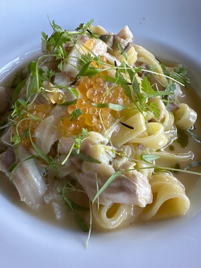 A close-up, high-angle shot shows a plate of pasta with fish and roe, likely from a restaurant in Tremezzina, Italy. The dish features wide, flat pasta, tender white fish flakes, and vibrant orange roe, all coated in a light, creamy sauce. Fresh green microgreens are scattered generously over the top, adding a touch of color and texture. The white ceramic plate is visible around the edges, suggesting an elegant presentation. The lighting is natural and soft, indicating an indoor setting with ample daylight. The focus is sharp on the food, highlighting its textures and colors, with a slight blur in the background.