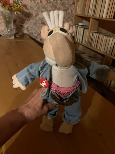 A stuffed toy, possibly a character from a video game, is held by a person's hand. The toy is dressed in denim overalls and a white shirt. It has a light brown head with black eyes and white ears that resemble tufts of fur. A red button with a white "M" is pinned to the toy's overalls. The background shows shelves filled with books and a speckled floor. The lighting suggests it is indoors, possibly in the evening. The hand holding the toy appears to be an adult's. The overall scene is casual and domestic.