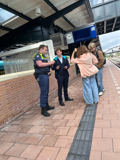 The scene depicts a public train platform at Groningen Hoofdstation in Groningen, Netherlands. The platform is paved with reddish-brown rectangular tiles, featuring a dark grey tactile warning strip. Above, a robust structure of black riveted iron beams supports a white-paneled ceiling with skylights and a cylindrical light fixture. To the left, a low brick wall separates the platform from an unseen area.
Four individuals are present. On the left, a male municipal enforcement officer (Handhaver) of medium build with light brown hair wears a dark blue uniform, a bright yellow reflective vest with a security badge, and a utility belt equipped with a holster, communication device, and a set of keys. He is looking down at an item held in his hands. Next to him, a male train staff member, with light brown wavy hair, is dressed in a dark blue suit with a light blue shirt, covered by a teal outer vest featuring an obscured logo. He holds a black smartphone or device in his left hand, engaging in an apparent interaction with the officer.
Further right, a female civilian with long brown hair, partially turned away from the camera, wears a light pink long-sleeved shirt and wide-leg light blue jeans, adjusting her hair with her right hand. Behind her stands a male civilian with blonde curly hair, dressed in a brown jacket, blue jeans, and white sneakers, carrying a dark backpack. Both are facing towards the train tracks visible on the far right, which include an overhead gantry and signal lights.
An overhead digital display board shows "2" for the platform number, a clock indicating approximately 10:40, and the text "Near train: 10:48 RS7 Veendam," indicating the next train service and destination. A small white sign with a toilet symbol is also visible near the display.