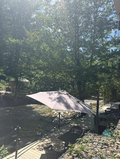 An outdoor seating area with tables and chairs is situated beside a shallow, rocky stream in a wooded area. A large, light-colored umbrella is open, casting shadows on the wooden deck and some of the furniture. The stream flows over rocks and gravel, with lush green trees and foliage surrounding the scene. In the background, there are two small structures with canvas roofs, possibly gazebos or seating areas, and more dense vegetation. The sky is visible through the trees, indicating a bright, sunny day. No people are present in the image. The location is identified as Shirokaya Schel, Russia.