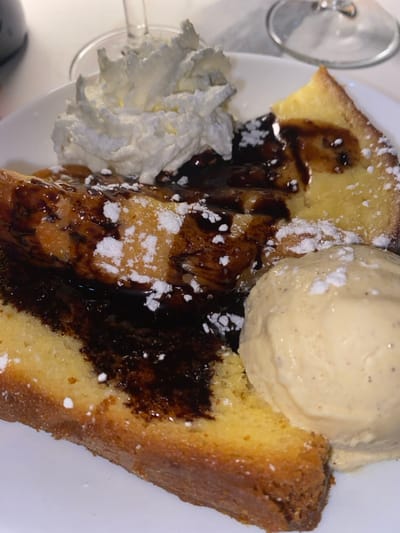 The image presents a high-angle, close-up view of a plated dessert, observed within an indoor dining setting, specifically identified as being in Cucuron, France. The dessert rests on a white, circular ceramic plate. The primary components include two substantial slices of a light-colored, golden-brown cake or bread, possibly pound cake or brioche. Centrally located on top of these slices is a rectangular item with a distinctive grid pattern, resembling a waffle or French toast, thoroughly drenched in a glossy, dark brown liquid, consistent with chocolate syrup. A large, irregularly mounded dollop of white whipped cream is positioned towards the upper left side of the plate. To the right, a scoop of pale yellow ice cream, displaying minute dark speckles, suggestive of vanilla bean, rests against one of the cake slices. A fine dusting of white powdered sugar is scattered over the chocolate-covered waffle-like item and the cake slices. In the blurred background, the upper portions of clear glass stemware are visible, indicating the presence of at least two drinking glasses on a light-colored surface. No individuals are present in the scene, which is static.