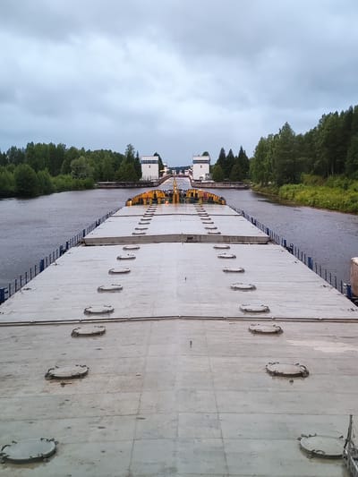 An overhead perspective shot, looking down the deck of a large, flat-decked barge. The barge is gray and appears to be constructed of concrete. Multiple circular, metal hatches are visible, evenly spaced across the deck. The barge is on a wide, calm river. On either side of the river, dense green forests line the banks. In the distance, a canal lock system is visible, featuring white control towers with the number "5" clearly displayed on one of them. The sky is overcast and a muted gray. No people are clearly visible in the frame. The location is Alekseevskoe, Russia.