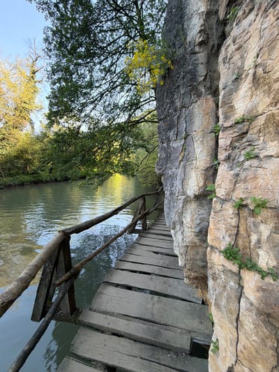 A landscape scene depicts a narrow river or canal with a wooden plank walkway running alongside it. The waterway reflects light and trees. Trees line the bank opposite the walkway. Above the walkway is a rocky overhang. The photo was taken in Petrevene, Bulgaria. There are no people visible in the frame.