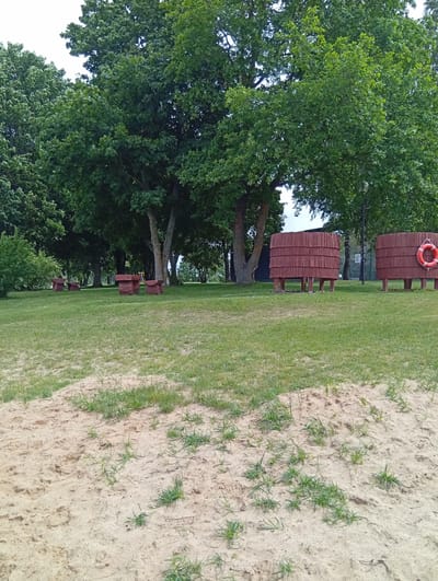 The image depicts an outdoor scene with a sandy foreground and a grassy area beyond. In the grassy area, there are several wooden structures that appear to be benches or tables with attached seating. Further in the background, two large, cylindrical wooden structures are visible, one of which has an orange life preserver attached to it. These structures are elevated on wooden legs and are situated amidst lush green trees. The environment suggests a park or recreational area, potentially near a body of water given the presence of the life preserver. There are no people or animals visible in the frame. The overall impression is one of a calm, natural setting.