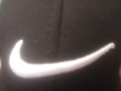 The image shows a close-up, blurry view of a white Nike swoosh logo on a black fabric surface. The logo appears to be slightly raised or textured. The lighting in the image is harsh, causing bright reflections on the logo, making it difficult to discern the exact texture and details of the fabric. The background is a dark, out-of-focus black. There is no information present in the image that would suggest a location in Valkosel, Bulgaria, or any other specific geographical context.