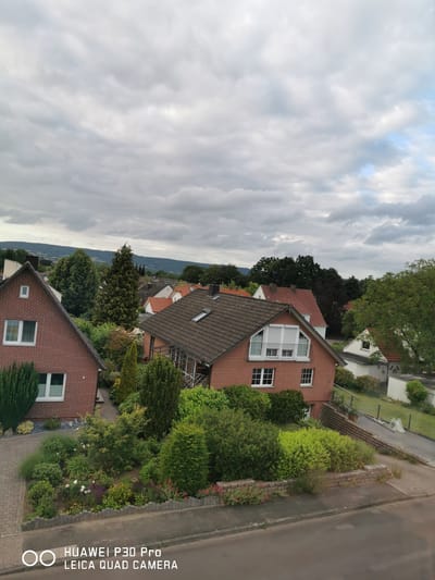 This is an eye-level, outdoor, full shot photograph taken on a cloudy day from an elevated vantage point looking down upon a residential area. The scene depicts a collection of houses with red-tiled roofs, interspersed with abundant green trees and manicured gardens. In the foreground, a street runs horizontally across the bottom of the frame, with a brick walkway and a lush garden leading up to a red brick house on the left. Further into the scene, more houses are visible, along with a winding road. In the background, a range of hills can be seen under the overcast sky. The image also includes a watermark at the bottom which reads "HUAWEI P30 Pro LEICA QUAD CAMERA". This scene is located in the city of Bad Oeynhausen, Germany, likely a quiet suburban or semi-rural neighborhood given the presence of detached houses, gardens, and surrounding greenery.