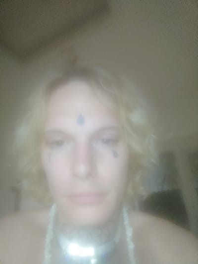 This image is a blurry, overexposed portrait of an individual, likely captured indoors in the city of Lichfield, United Kingdom. The subject, an adult with light skin and wavy, shoulder-length blonde hair, is depicted from the upper torso upwards. Distinctive features include a small ring piercing in the left nostril, a dark tear-drop shaped marking below the outer corner of each eye, and a similar dark, oval or tear-drop shaped marking centered on the forehead. The neck displays dark, intricate patterns, consistent with a tattoo. The individual wears a multi-strand light-colored beaded necklace and a wide, reflective, metallic choker, which is severely overexposed by a bright glare in the lower center of the frame. The subject's eyes are cast downwards. The background is uniformly light-toned, indistinct, and heavily blurred, providing no discernible environmental details. No specific actions or interactions beyond posing for the photograph are visible.