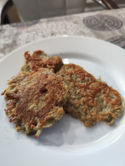 A close-up shot shows three golden-brown potato pancakes on a white plate, likely taken indoors in a kitchen or dining area in the city of Moskovskiy, Russia. The pancakes are irregular in shape and appear to be crispy on the edges. A blurred background suggests a table setting with a patterned tablecloth.