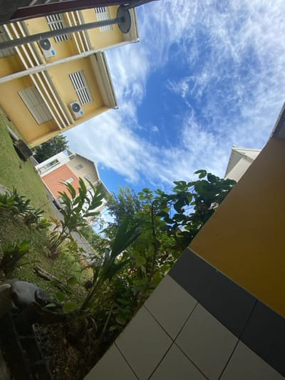 This image captures an upward-angled view of an outdoor residential or commercial environment in Capesterre-Belle-Eau, France. The scene is illuminated by natural daylight under a vibrant blue sky exhibiting scattered, wispy white clouds.
In the upper-left quadrant, a two-story building, painted a light yellow, features white horizontal louvered windows on both levels. Each floor is equipped with an external air conditioning compressor unit mounted below its respective window. A dark gray or black vertical pole, possibly structural, extends from the bottom left of the frame, partially obscuring the yellow building and culminating in a circular component at its apex.
The ground level to the left presents a grassy incline populated by diverse green tropical vegetation, including broad-leafed plants and shrubs. Further in the mid-ground, slightly left of center, a distinct multi-story building with an orange lower facade and a lighter, possibly white or beige, upper section with a sloped roof is visible. A road runs in front of this building, with a dark-colored vehicle partially visible on it.
The lower right quadrant of the image reveals a building corner, characterized by a yellow upper section, a horizontal dark gray band, and a tiled base composed of white square tiles. Dense green foliage and tree canopy elements occupy the central and right portions of the lower and mid-ground, partially obscuring the distant horizon and another white-roofed building on the right. No people are visible.