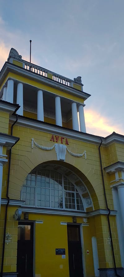 The image shows the facade of a yellow building with white columns and neoclassical architectural elements. The name "ЛУГА" (Luga) is written in red Cyrillic letters above a large arched window. A small clock is mounted on the wall to the left of a dark door, and a sign with Cyrillic text is visible near another dark door on the right. The sky in the background displays a mix of blue and orange clouds, suggesting it might be sunset or sunrise. The building appears to be the Luga railway station in Luga, Russia.