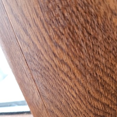 A close-up, angled photograph depicts two adjacent panels of a wood-grain patterned surface, likely laminate flooring or wall paneling, in an interior setting in Bachevo, Bulgaria. The panels exhibit a rich medium-brown coloration with darker, irregular linear patterns indicative of wood grain. A precise, vertically oriented seam, angled slightly from the upper left to the lower left, separates the two panels. No people are visible. In the lower-left foreground, a blurred, bright white area with a faint horizontal dark line beneath it is visible, suggesting an out-of-focus window or an illuminated opening. No actions or explicit interactions beyond the structural joining of the panels are discernible.