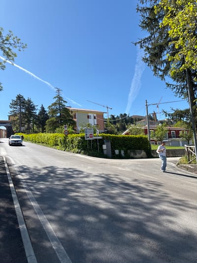 This image captures a bright, sunny day in what appears to be a semi-urban or residential area, consistent with Camerino, Italy. An asphalt road curves gently to the left, with pronounced, dappled shadows from unseen trees stretching across its surface, indicating strong sunlight.

On the right side of the road, a person stands facing away from the camera, looking down the street. They are wearing a light-colored t-shirt and blue jeans. Behind them is a tall, well-maintained green hedge, and beyond that, various buildings, including a structure with a red-tiled roof and other multi-story buildings with different facades. In the background, at least two large construction cranes are visible, suggesting ongoing building or reconstruction activities.

Further down the road on the left, a small, dark-colored compact car (possibly electric) is visible. The roadside is lined with numerous green trees and lush foliage. The sky above is a clear, vibrant blue, traversed by two distinct white contrails from airplanes.

Several road signs are visible on the right. A circular sign with a red border prominently displays "30," indicating a speed limit of 30 km/h. Above it, another sign points towards "Centro" (center) and includes a parking symbol. Partially visible text near the small car on the left reads "ROONADRIATICO," which could be a street name or part of a business name. The scene conveys a calm, everyday atmosphere under clear skies.
