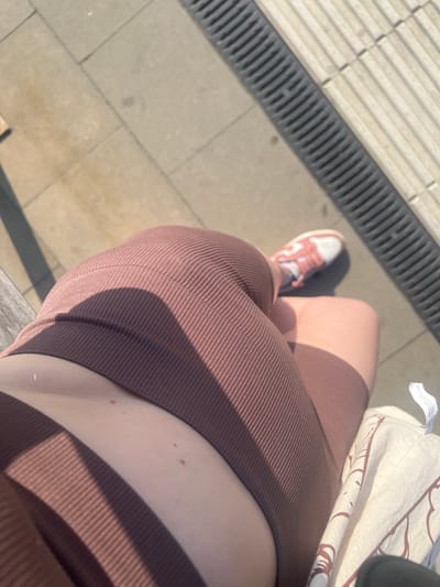 The image shows a close-up, top-down view of a person's legs and torso, sitting outdoors on what appears to be a sunny day. The person is wearing brown ribbed athletic shorts and a white and pink sneaker. The background consists of light-colored paving stones and a dark grated drain. The scene is likely located in Skawina, Poland, given the context provided. There are no other people or distinct architectural features visible. The lighting suggests direct sunlight, casting shadows across the scene.