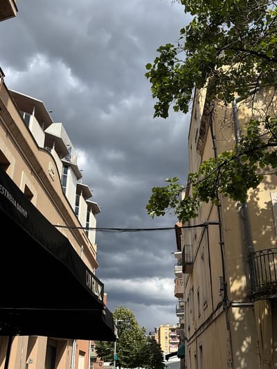 The image is oriented vertically, showing a street scene in Granollers, Spain, under a cloudy sky. The perspective is from the street level looking up. Buildings line both sides of the street, with varying architectural styles. The building on the left is tan, with stepped architectural elements. A large black awning with the text "ESTRELLA DAMM" is visible on this building. The buildings on the right are lighter in color, and traditional in style. Green foliage from trees overhangs the scene from the top left and right, partially obscuring the buildings. A vertical power line runs between the buildings. The sky is filled with dark grey clouds.