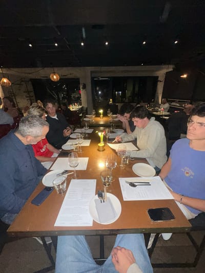A group of people are sitting at a long table in a dimly lit restaurant. The table is set with plates, silverware, wine glasses, and menus. A few decorative centerpieces sit on the table. People are dressed casually. Some are looking at menus and others are talking. A dark-colored smartphone sits at the top of the table. Overhead, decorative lights hang from the ceiling. Outside, city lights are visible through a window.