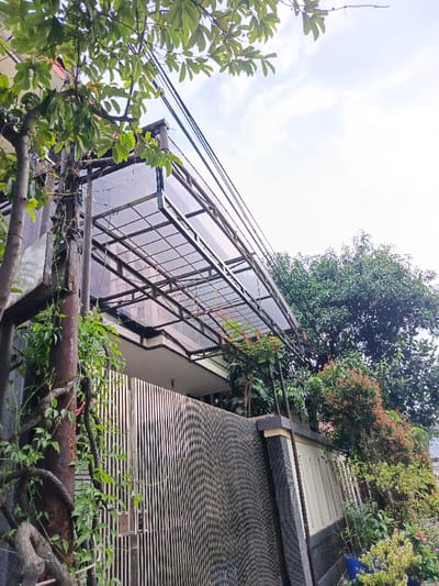 A low-angle shot depicts a residential property with a covered, translucent patio roof. A metal fence with vertical slats lines the perimeter. Lush greenery, including trees and climbing vines, partially obscures the structure. An utility pole with wires runs across the top of the image. The sky is bright with a few scattered clouds. The overall scene conveys a sense of nature intertwined with urban architecture.