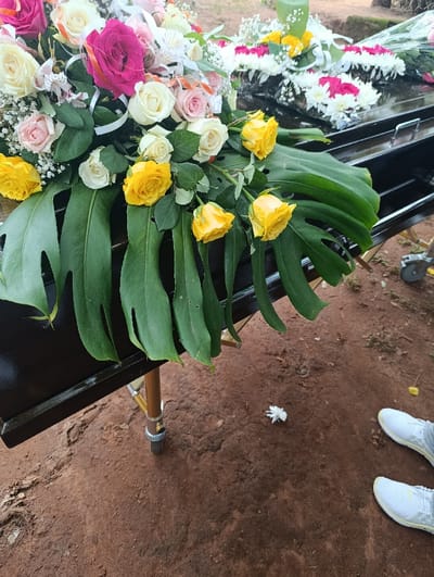 The scene depicts a black polished casket situated outdoors on reddish-brown earth in Muthetheni ward, Kenya. A large, elaborate floral arrangement adorns the casket's upper surface, predominantly featuring white, pink, yellow, and fuchsia roses, accented by white alstroemeria, baby's breath, and scattered orange and white ribbons. Several large green monstera leaves are laid beneath the main arrangement, with additional yellow roses placed individually along their length. Further down the casket, another floral arrangement containing white and pink chrysanthemums with yellow accents is visible. The casket rests on a metallic stand, with a gold-colored leg and a wheel partially visible. On the ground, a single fallen white flower and small debris are present. The lower legs and feet of an individual, wearing white athletic shoes, are visible in the bottom right foreground.