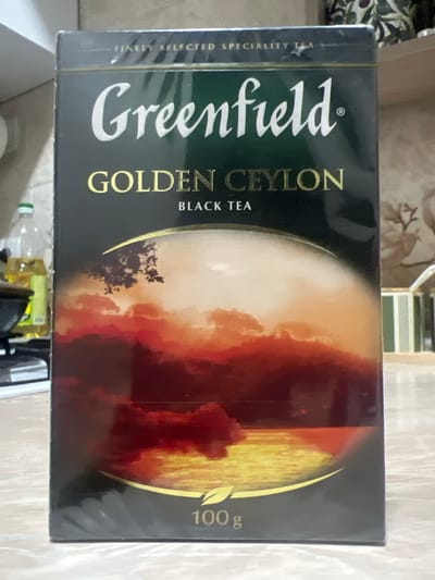 A box of Greenfield Golden Ceylon black tea is centered in the frame, standing upright on a light-colored, textured surface. The box has a dark background with a gradient that transitions from black at the top to a reddish-brown at the bottom. The brand name "Greenfield" is prominently displayed in large white cursive font. Below it, in gold lettering, are the words "GOLDEN CEYLON," and beneath that, "BLACK TEA." A scenic image is depicted on the lower half of the box, featuring a dusky sky with fiery clouds and a bright, reflective body of water. A stylized golden leaf logo is visible near the bottom, with the text "100 g" also present. The box appears to be sealed in plastic wrap, indicated by subtle reflections and creases on its surface. In the background, to the left, a partially visible bottle with a yellow liquid and a white cap can be seen, along with a portion of a stovetop and kitchen counter. To the right, out-of-focus elements suggest a shelf or cabinet with decorative items. The lighting is moderate, casting soft shadows.