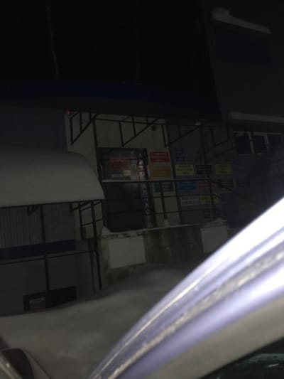 Captured from a skewed perspective, the image reveals a building façade at night. The building exhibits a metal frame adorned with various advertisements and notices. A storefront with signage in a Cyrillic script is visible. The scene is dimly lit, with the darkness amplifying the visual impact of the lit advertisements. Part of a car appears in the foreground.