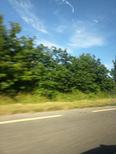 The image captures a blurred, motion-filled view from a moving vehicle, likely on a road near Killarney, Ireland. The scene is dominated by lush green trees and vegetation on the left side, suggesting a rural or roadside setting. A clear blue sky with wispy white clouds is visible above the trees. A grey asphalt road with a distinct yellow line marking its edge runs across the lower portion of the image, indicating a paved surface. The motion blur suggests the photograph was taken while the vehicle was in transit, possibly on a scenic route or highway in the vicinity of Killarney, known for its natural beauty. There are no visible people or specific man-made structures within the frame, focusing the composition on the natural landscape and the road.