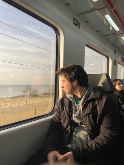 Inside a train, a young man sits near the window, gazing out. He wears a dark jacket over a gray hoodie, with white earbuds plugged in. Outside, a view of a sandy beach and lake is visible. In the background, another person is seated, leaning back and appearing to sleep. The train interior is white and has large windows. A reflection of the man's face can be seen in the window.