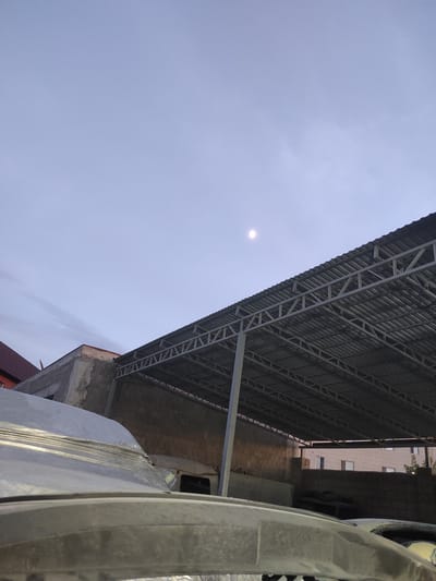 This media file shows an outdoor scene under a pale, overcast sky. The image is oriented vertically, with the sky occupying the upper left portion. A large, angled metal roof structure with a corrugated metal surface and exposed metal beams dominates the right and upper right of the frame. The roof appears to be part of an industrial or construction site, with visible cinder block walls beneath it and some scaffolding or support beams.
To the right of the roof structure, a building with light-colored walls and a few rectangular windows is visible in the background. There is a hint of greenery on a tree branch near the top of this building. In the foreground, on the far right side of the image, a portion of a large, textured object, possibly a vehicle or a piece of construction equipment, is visible. Its surface is a dull gray and appears somewhat weathered.
The overall impression is one of a construction or industrial area, possibly during the early morning or late evening given the muted light and the presence of what looks like a faint moon or planet in the sky. There is no visible text in the image. The scene is devoid of people and focuses on the architectural elements and the sky.
