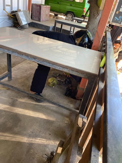 A person wearing a black shirt, black pants, and a black helmet is lying face down under a tiled table in Santan, Indonesia. The person's legs are extended towards the right of the frame, with their right leg partially obscured by the table. A round, green fruit, possibly a jackfruit or durian, rests on the ground near the person's legs. The environment appears to be an outdoor seating area with a concrete floor and a wooden railing. In the background, a green vehicle is parked, and a building with white vertical siding is visible. A young boy is partially visible to the right of the frame, looking towards the camera.
