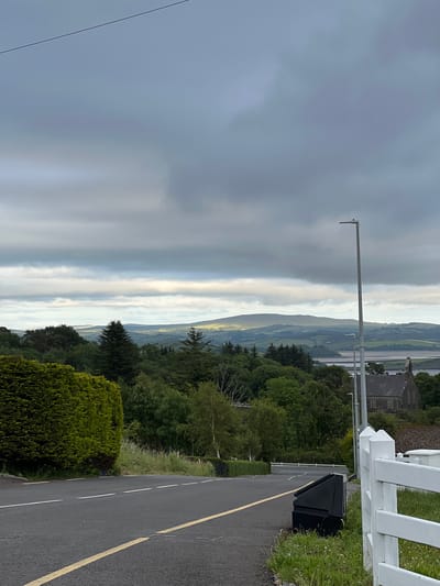 The image depicts a scenic landscape in Mountcharles, Ireland, with a road in the foreground. The road is asphalt with a yellow line marking the edge and a white line dividing the lanes. To the right of the road is a white wooden fence and a black trash bin. To the left of the road, there is a grassy area with some wildflowers. Beyond the road, a dense forest of green trees stretches out. In the distance, rolling hills are visible, some of which are covered in vegetation. The sky is overcast with streaks of light breaking through the clouds, suggesting either early morning or late evening. The overall mood is serene and peaceful, capturing the natural beauty of the Irish countryside. There are no people visible in the image, and no text is present. The lighting and the overcast sky indicate a diffused light, possibly during a cloudy day or at dawn/dusk.