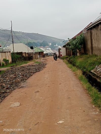An overcast, medium shot captures a dirt road in Ibo, Nigeria, with a motorcycle in motion. The road is predominantly reddish-brown dirt, with patches of loose stones and some larger flat rocks scattered on the left side. To the right, a grassy verge transitions into a drainage ditch. The surrounding environment includes low-rise buildings with corrugated iron roofs and concrete walls, some featuring barbed wire fences. Dense green vegetation grows along the roadside and in front of some of the buildings. In the background, a sloping, rocky hill dominates the skyline under a hazy, grey sky. The date stamp "2026/06/13 09:10" is visible in the bottom left corner.