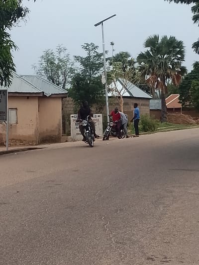 The image captures a street scene in what appears to be Biliri, Nigeria. A paved road stretches into the distance. Along the roadside, there are small, single-story buildings with corrugated metal roofs. Three men stand near two motorcycles. One man is seated on a motorcycle, actively riding. Another man wearing a red shirt is leaning over the front of another motorcycle. A third man in a blue shirt is standing adjacent to them. In the background, palm trees are visible, along with a light pole. A sign can be seen near one of the buildings.