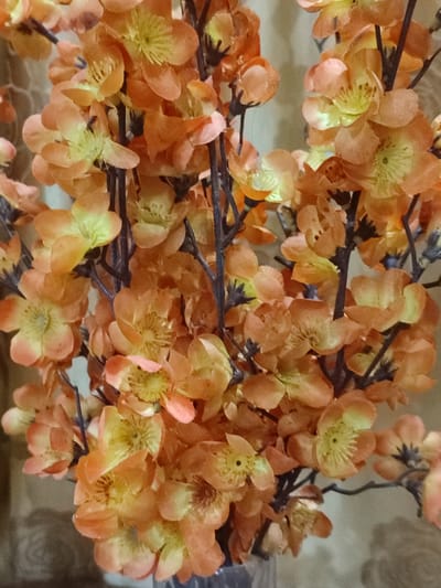 This is a close-up, vertical, medium shot of artificial orange and yellow flowers. The flowers are densely packed on dark brown stems, creating a lush and vibrant bouquet. Each flower has multiple delicate petals and a detailed center with yellow stamens. The lighting is somewhat dim and diffused, with hints of light catching the edges of the petals. The background is out of focus, showing a textured, light-brown surface that could be a curtain or wall hanging, possibly with a subtle pattern. The overall impression is one of artificial beauty and a decorative arrangement, likely indoors. There are no people, specific activities, or identifiable text visible. The location context of Blang Cut, Indonesia, does not provide any direct visual cues within the image itself.