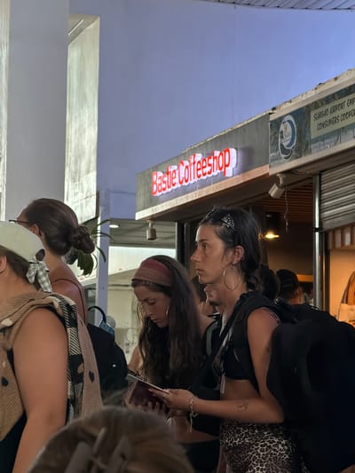 The image shows three women standing on a street outside "Bastie Coffeeshop" in Del Carmen, Philippines. The cafe sign is red. One woman wears hoop earrings, a headband, and has a tattoo on her arm. Another wears a red headband, and the third is wearing a plaid scarf, a hat, and has their hair up in a bun. The lighting suggests it is daytime. A sign indicates this is the location of the Siargao Airport Employees Consumers Cooperative.