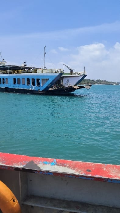 The image depicts an aquatic scene in the Ganjoni/Shimanzi ward, Mombasa, Kenya. A large, multi-decked blue and white vehicular ferry, bearing the hull number "JA 006" on its side, occupies the midground. The ferry's bow ramp is visible in a raised position, and several individuals are discernible on its upper deck. Masts and other maritime equipment are present on the upper structure. The ferry floats on calm, turquoise-blue water. In the distance to the right, a low-lying landmass covered in green vegetation is visible. The sky is partly cloudy blue. The foreground shows a weathered red and white railing with chipped paint, from which the photograph appears to have been taken, and a partial orange object, possibly a lifebuoy or fender, on the lower left. The ferry appears to be in motion or stationary in open water, not actively engaged in docking or loading procedures.