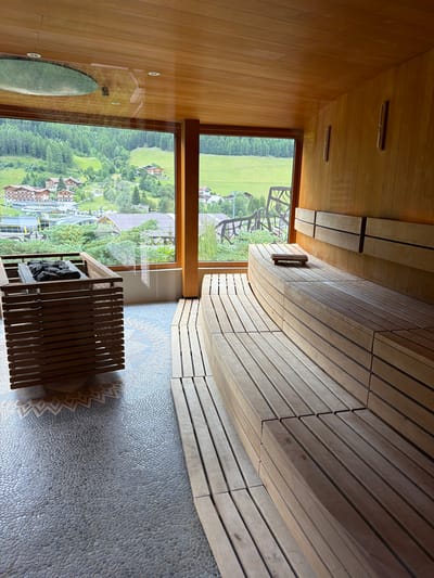 The image shows an interior view of a sauna with large windows overlooking a scenic mountainous landscape. The sauna features wooden benches with a curved design, a wooden structure containing heated stones, and a floor tiled with small pebbles in a mosaic pattern. Through the windows, a village with traditional houses and green hills covered with trees are visible. The architecture of the sauna suggests a modern design integrated with natural materials. The lighting is warm and ambient, typical of a sauna environment. The scene is static, with no people or actions depicted, creating a serene and tranquil atmosphere. The view from the sauna suggests it is located in a mountainous region, likely a resort or spa, possibly in Mühlbach - Rio di Pusteria, Italy, given the architectural style and landscape.