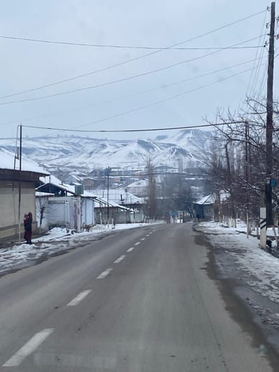 This image depicts a snow-covered street scene in Norayburi, Uzbekistan, under an overcast sky. A two-lane asphalt road with white dashed center markings extends into the distance, partially cleared of snow, but flanked by snow and slush on its shoulders. On the left side of the road, a person in a dark red, full-length garment stands near a building, holding what appears to be a swaddled infant or small child. Buildings with snow-covered roofs line both sides of the road, receding into the background. Bare deciduous trees with snow on their branches are visible along the right side of the road and in the middle distance. Utility poles, both wooden and metal, with multiple overhead power lines, are present on both sides of the road. In the background, snow-capped mountains dominate the horizon.