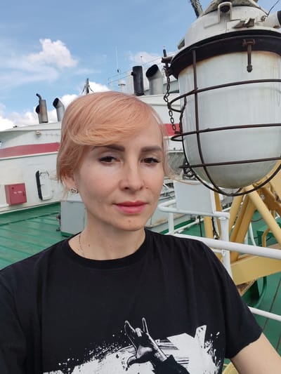 In a close-up, outdoor, selfie-style photograph taken on a ship, a woman with short, peachy-blonde hair is the primary subject. She is looking directly at the camera with a neutral expression. Her makeup includes visible eyeliner and mascara, and she is wearing a small, heart-shaped earring with clear stones in her left ear. She is also wearing a dark t-shirt with a graphic print.
The background features elements of the ship's deck, including green painted metal flooring and white railings. To the right of the woman, a large, white, globe-shaped light fixture with a protective metal cage is prominently visible. In the distance, part of the ship's superstructure is visible, painted white and red, with dark-colored smokestacks. The sky above is partly cloudy. The image does not provide any specific visual cues to suggest the location is the city of Белоусово, Russia.